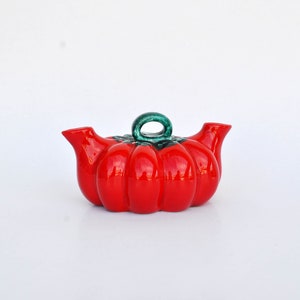 Può includere: Una teiera di ceramica rossa a forma di pomodoro con gambo e manico verdi.