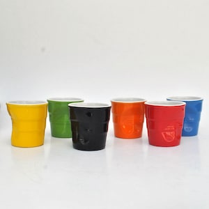Può includere: Un set di sei tazze in ceramica colorate: giallo, verde, nero, arancione, rosso e blu. Le tazze hanno un design unico e strutturato e sono disposte su una superficie bianca. Una scatola nera è visibile sullo sfondo.