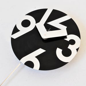 Op de afbeelding: Een ronde wandklok met een zwarte wijzerplaat en grote, witte, gestileerde cijfers. De klok heeft twee dikke, witte, hoekige wijzers die naar rechtsboven wijzen. Een dunne, rechte, witte pendelstang steekt vanuit het midden naar beneden.