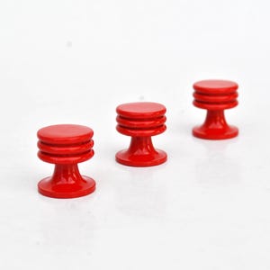 May include: Three small, red, wooden knobs with a round top and a tiered, cylindrical body. The knobs are evenly spaced against a white background. The knobs are likely for furniture or decorative purposes.