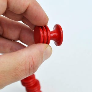 May include: A bright red wooden knob, held in a hand, against a white background. The knob has a rounded top and a cylindrical body with a series of stacked, circular elements. The knob is part of a longer, matching red wooden piece.