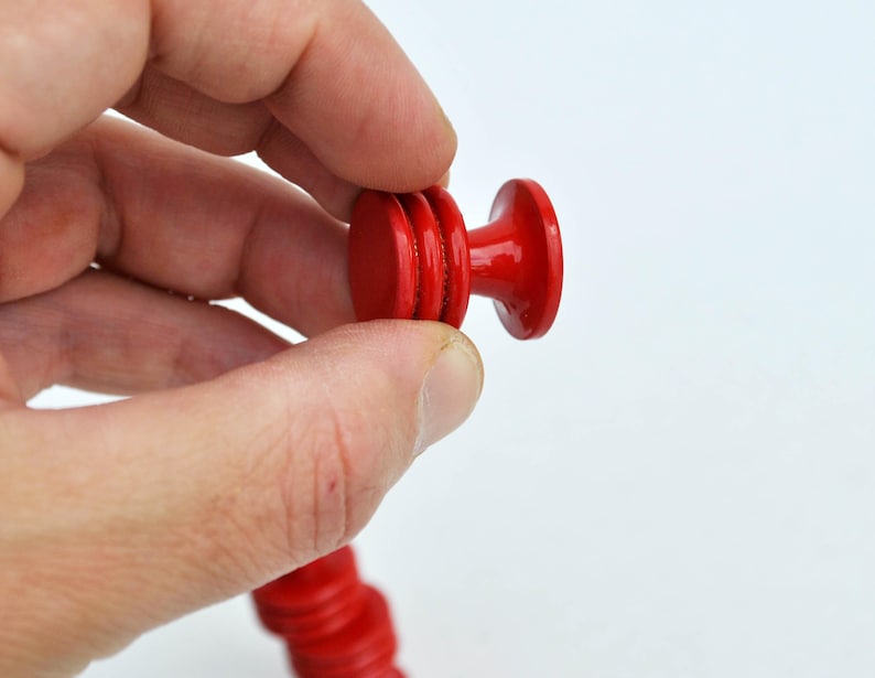 May include: A bright red wooden knob, held in a hand, against a white background. The knob has a rounded top and a cylindrical body with a series of stacked, circular elements. The knob is part of a longer, matching red wooden piece.