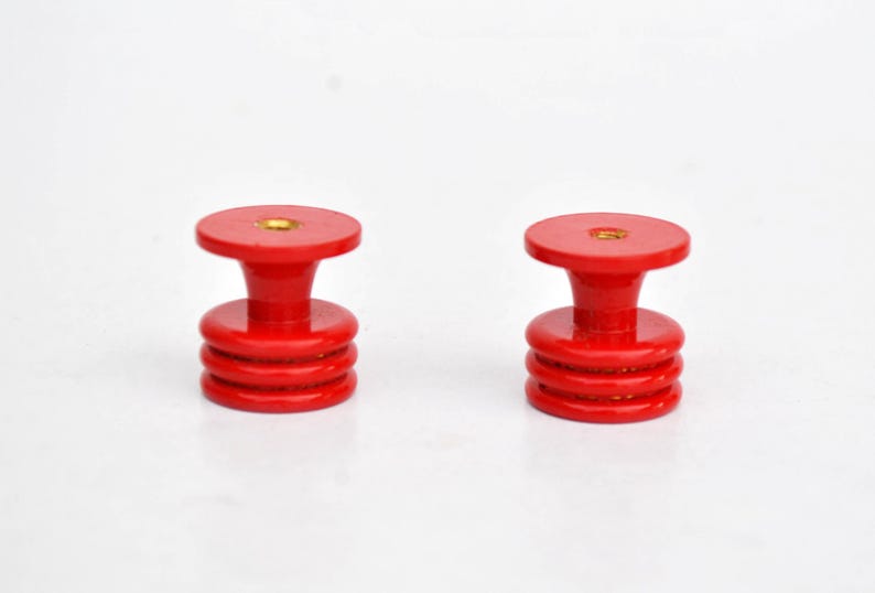 May include: Two red cabinet knobs with a stacked, cylindrical design. Each knob has a flat, circular top and a flared base. The knobs are made of a glossy, solid red material and are set against a white background.