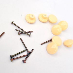 May include: A collection of pale yellow cabinet knobs and screws. The knobs are round with a flat base and a slightly domed top. The screws are bronze-colored with a slotted head.