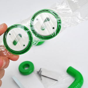 May include: Two green and white circular objects with a black border are held in a clear plastic bag. Additional green components and hardware are visible on a white surface. The objects appear to be parts of a larger product.