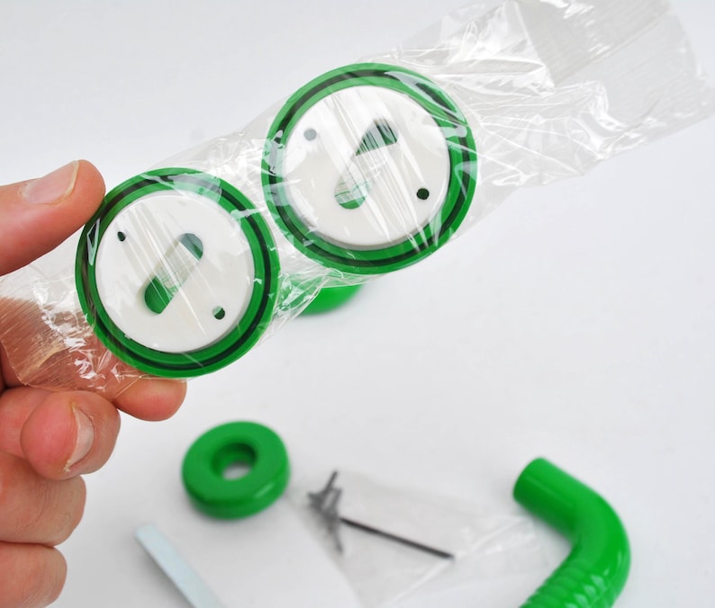 May include: Two green and white circular objects with a black border are held in a clear plastic bag. Additional green components and hardware are visible on a white surface. The objects appear to be parts of a larger product.