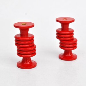 May include: Two red wooden knobs with a stacked disc design. Each knob has a round top and base, connected by a series of smaller, circular discs. The knobs are painted a vibrant red color and sit against a white background.