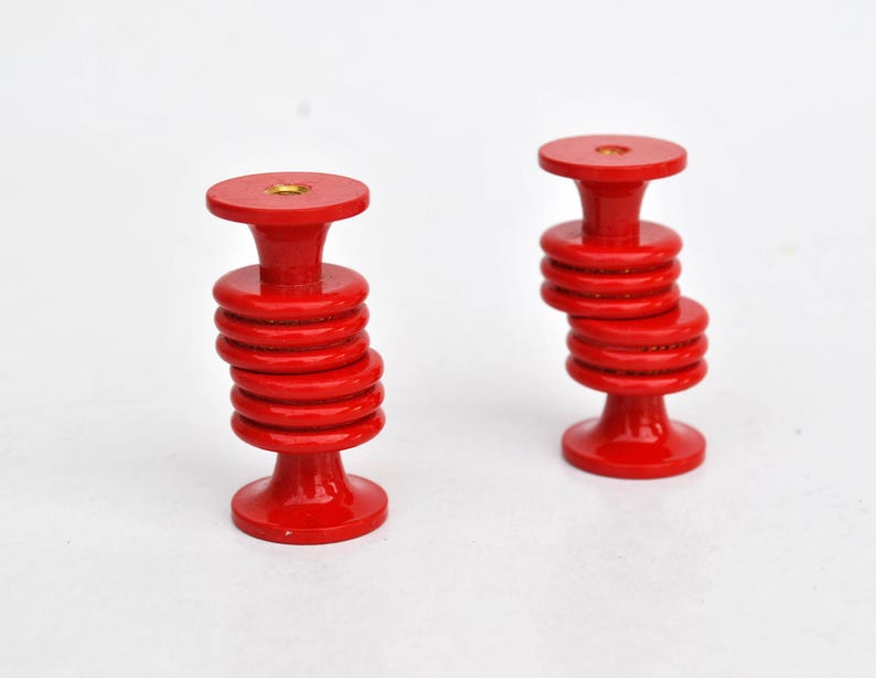 May include: Two red wooden knobs with a stacked disc design. Each knob has a round top and base, connected by a series of smaller, circular discs. The knobs are painted a vibrant red color and sit against a white background.