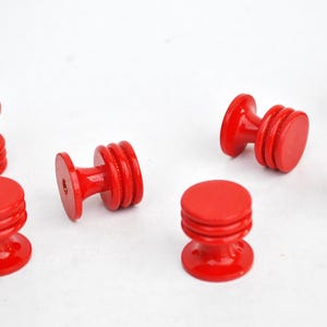 May include: Several bright red wooden cabinet knobs are arranged on a white surface. Each knob has a round top and base, connected by a ridged, cylindrical body. The knobs are approximately 1.5 inches in diameter.