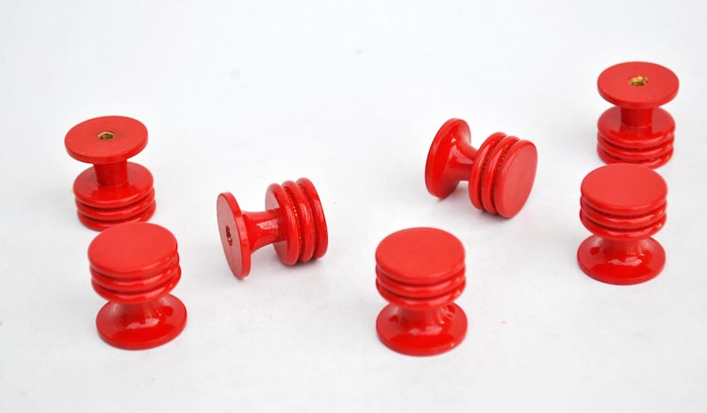 May include: Several bright red wooden cabinet knobs are arranged on a white surface. Each knob has a round top and base, connected by a ridged, cylindrical body. The knobs are approximately 1.5 inches in diameter.