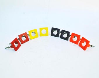 May include: A set of eight square cabinet knobs arranged in an arc. The knobs feature a central circular cutout and come in a variety of solid colors including red, yellow, black, and orange. Each knob has a metal screw post extending from the back.