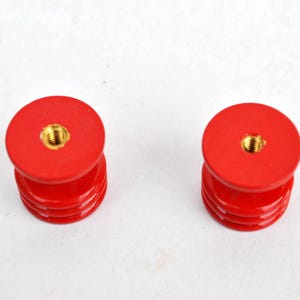 May include: Two red, cylindrical objects with a threaded brass insert on top. The objects have a stacked design with multiple horizontal ridges. The background is plain white.