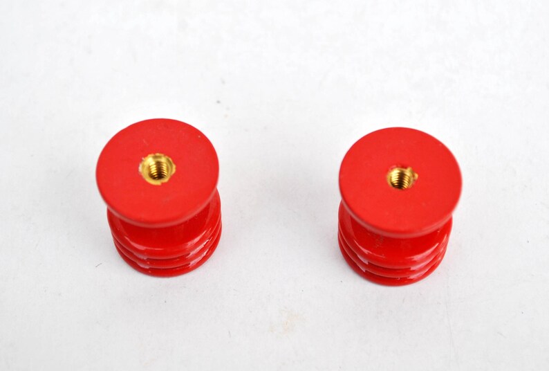 May include: Two red, cylindrical objects with a threaded brass insert on top. The objects have a stacked design with multiple horizontal ridges. The background is plain white.