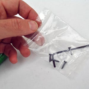 May include: A clear plastic bag containing several small black screws and a black Allen wrench. The items are held in a person's hand, with a green object visible in the lower left corner.