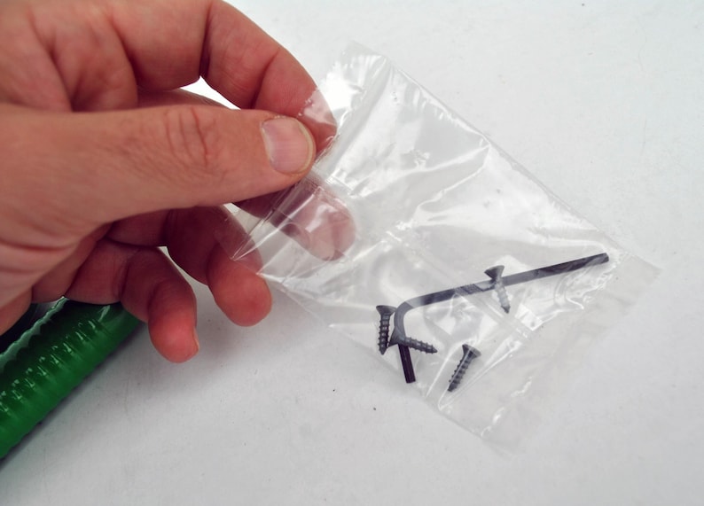 May include: A clear plastic bag containing several small black screws and a black Allen wrench. The items are held in a person's hand, with a green object visible in the lower left corner.