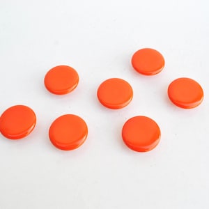 May include: Seven bright orange, round buttons are scattered on a white surface. The buttons have a glossy finish and a slightly raised centre. They are evenly spaced and appear to be made of plastic.