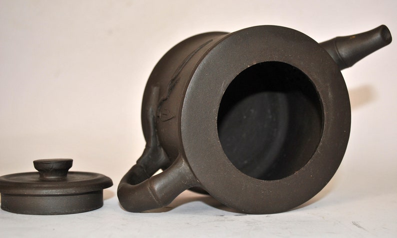 May include: A brown ceramic teapot with a lid and a spout. The teapot has a round body and a handle on the side. The teapot is sitting on a white surface.
