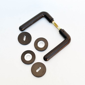 Puede incluir: Un juego de manillas de puerta marrones con una placa posterior cuadrada y cuatro rosetas redondas. Las rosetas tienen un centro blanco.