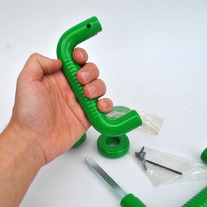 May include: A bright green, curved handle with a ridged grip, held in a hand. Additional green circular pieces, screws, and a metal tool are also visible, suggesting a construction or repair item.