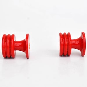 May include: Two bright red wooden knobs with a glossy finish. Each knob features a cylindrical body with three stacked rings and a flared base and top. The knobs are set against a white background.