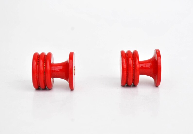 May include: Two bright red wooden knobs with a glossy finish. Each knob features a cylindrical body with three stacked rings and a flared base and top. The knobs are set against a white background.