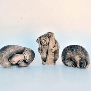 May include: Three ceramic animal figurines: a platypus curled up, a koala clinging to a tree trunk, and a spiky echidna. The figurines are all painted in shades of brown and gray.