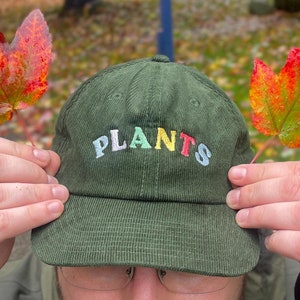 Può includere: Un berretto da baseball in velluto a coste verde con la parola "PLANTS" ricamata in lettere colorate. Il berretto è tenuto da una persona con le mani su entrambi i lati del berretto.