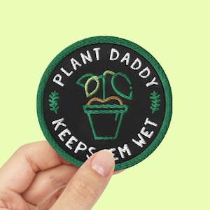 Può includere: Toppa ricamata verde con il testo "Plant Daddy Keeps 'Em Wet" e un'immagine di un cartone animato di una pianta in vaso.