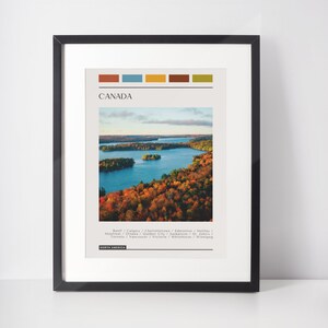 May include: A minimalist poster with a colorful stripe design at the top. The poster features a photograph of a lake surrounded by trees in autumn colors. The text "CANADA" is in a bold font at the top of the poster. Below the image, the text "Banff/Calgary/Charlottetown/Edmonton/Halifax/Montreal/Ottawa/Quebec City/Saskatoon/St. John's/Toronto/Vancouver/Victoria/Whitehorse/Winnipeg" is listed in a smaller font. The text "NORTH AMERICA" is at the bottom of the poster.