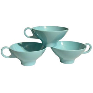 May include: Three light blue, vintage-style teacups are stacked in a triangular arrangement. Each cup has a rounded bowl, a flared base, and a curved handle. The cups are made of a solid, opaque material.