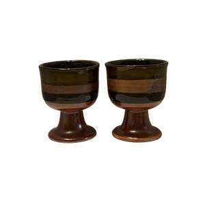 May include: Two ceramic goblets with a brown and black striped design. The goblets have a pedestal base and a rounded bowl. The color palette includes shades of brown, black, and a reddish-brown. The goblets are likely for serving beverages.