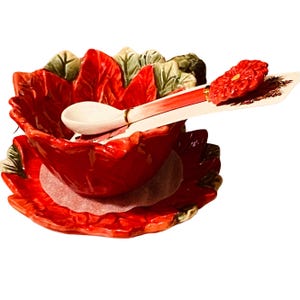 May include: A red ceramic serving bowl and saucer set, shaped like a flower, with green and white accents. A white spoon with red accents rests on the bowl. The set is designed to resemble a poinsettia.