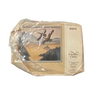 May include: A plastic-wrapped package containing a cross-stitch pattern for a nature scene. The pattern features two geese flying over a lake and forest. The package includes the text "The Creative Circle" and the product number "6184".