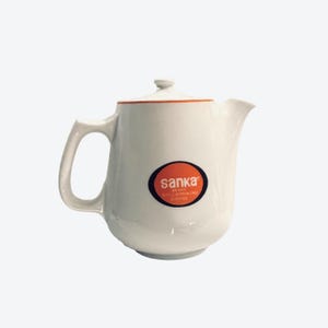 May include: White ceramic teapot with a lid and handle. The teapot has an orange trim and a circular logo that reads "sanka" in orange letters. The logo is on a black and red background. The teapot is designed for serving coffee.