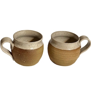 May include: Two handmade ceramic mugs. The mugs have a rounded shape with a light beige speckled top and a brown base. Each mug has a curved handle. The mugs are set against a white background.