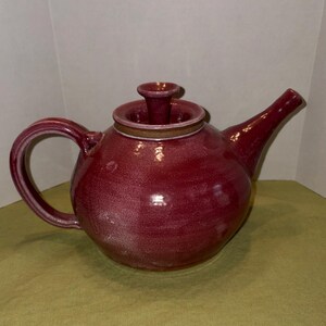 May include: A handcrafted, burgundy-colored ceramic teapot with a rounded body, curved handle, and a spout. The lid has a small, cylindrical knob. The teapot has a speckled finish and a light brown band around the rim.