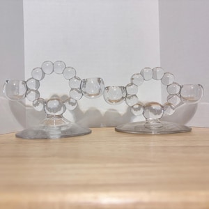 May include: Two clear glass candle holders. Each has a circular design of glass bubbles and three candle cups. The candle holders sit on a round base. The background is white and the surface is wood.