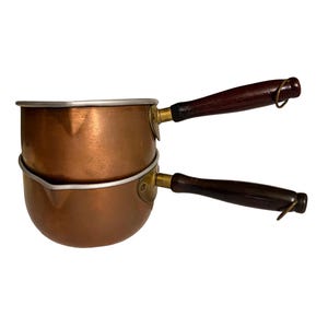 May include: Two copper saucepans stacked on top of each other. Each saucepan has a dark brown wooden handle and a metal rim. The handles are attached with brass-colored hardware. The copper has a warm, reddish-brown hue.