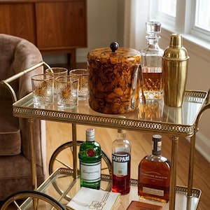 May include: A gold bar cart with a glass top, holding cocktail glasses, a cocktail shaker, and bottles of liquor. The cart has a decorative ice bucket and a decanter. The cart has wheels.