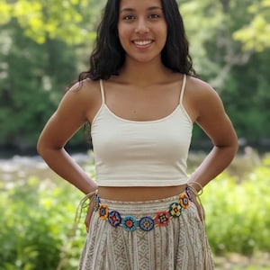 May include: A woman wearing a cream-colored crop top and a patterned skirt. A colorful beaded belt with circular designs in various colors is around the waist. The background is a blurred view of a natural outdoor setting.