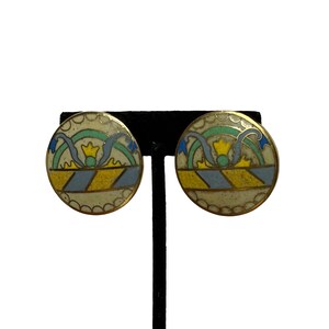 May include: A pair of round, gold-toned clip-on earrings. Each earring features a cream-colored background with a blue, yellow, and green design. The design includes a ribbon and crown motif. The earrings are displayed on a black stand.