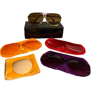 May include: Gold-framed aviator sunglasses with brown lenses sit atop a black box. Included are three pairs of colored lenses in orange, red, and purple cases, along with a yellow cleaning cloth and a lens. A vintage eyewear set.
