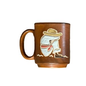 May include: Brown ceramic mug with a handle, featuring a relief design of a person in a sombrero and traditional clothing. The figure is depicted in white, red, and tan colors. The mug has a glossy finish.