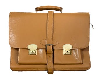May include: A tan leather briefcase with a top handle and a front flap secured by a buckle. The briefcase has two front pockets with gold-colored clasps. The leather has a textured appearance.