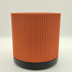 May include: A cylindrical planter featuring a vibrant orange upper section with a vertical ribbed texture and a solid black base. The container has a straight-sided profile and a smooth, flat rim, providing a clean and functional design for holding plants.