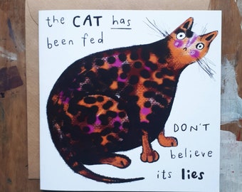 May include: A white card with a black and orange cat with pink spots. The text on the card reads "the CAT has been fed DON'T believe its lies".