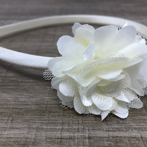 Puede incluir: Una diadema blanca con un adorno de flor de tela blanca. La flor está hecha de varias capas de tela y tiene un aspecto delicado y femenino.