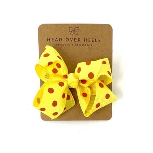 May include: Yellow hair bow with red polka dots. The bow is on a brown card with the text "Head Over Heels Unique Hair Accessories".
