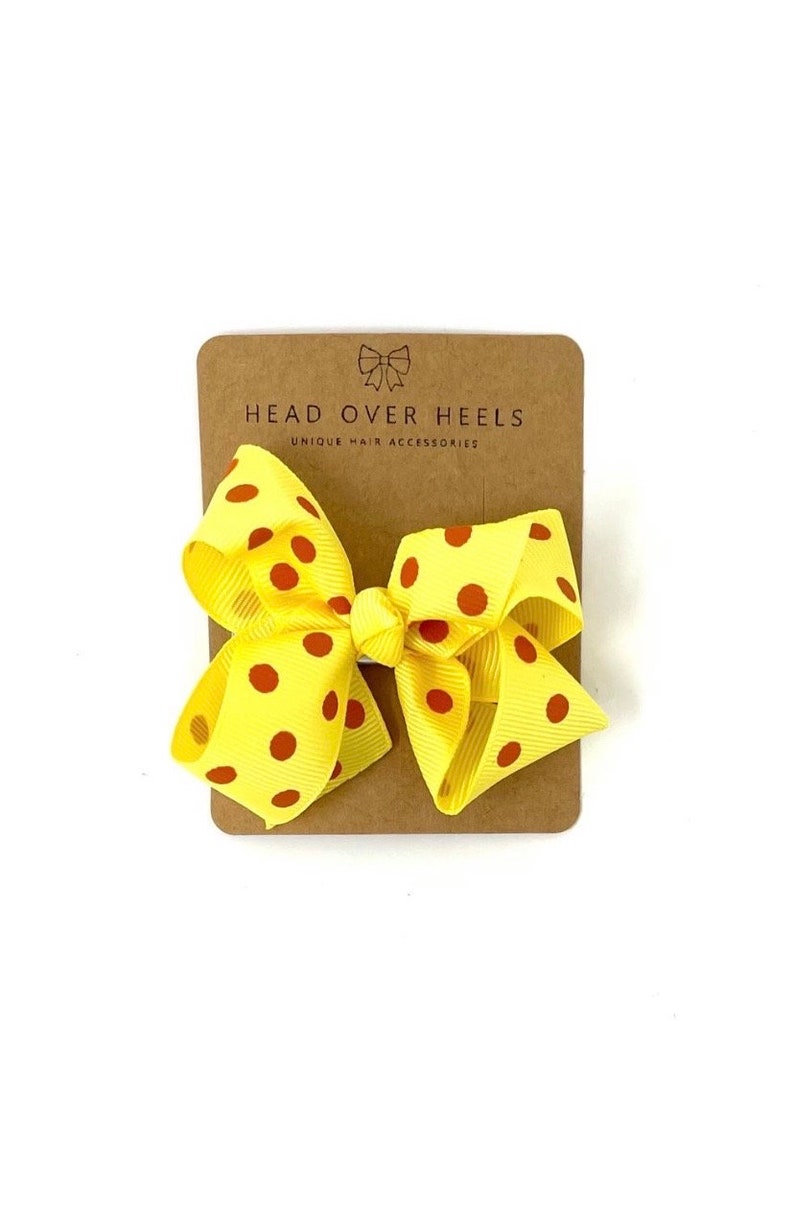 May include: Yellow hair bow with red polka dots. The bow is on a brown card with the text "Head Over Heels Unique Hair Accessories".