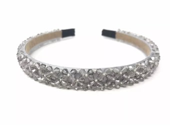 May include: A silver headband with a sparkling, crystal-like embellishment. The headband is covered in small, clear, faceted beads, creating a shimmering effect. The band itself is a light gray color with black ends.